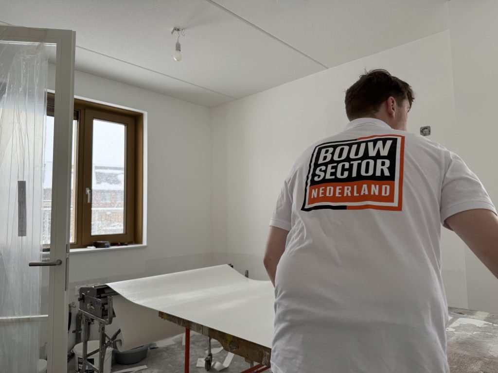 Behanger van Bouwsector Nederland bij de plaktafel met een baan behang in een lege kamer