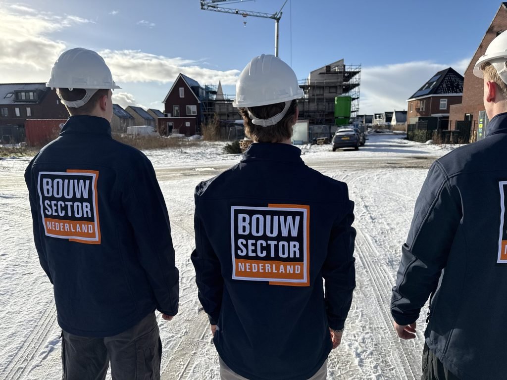 Drie mannen in werkkleding met logo op de rug voor een rij huizen in aanbouw onder een blauwe lucht